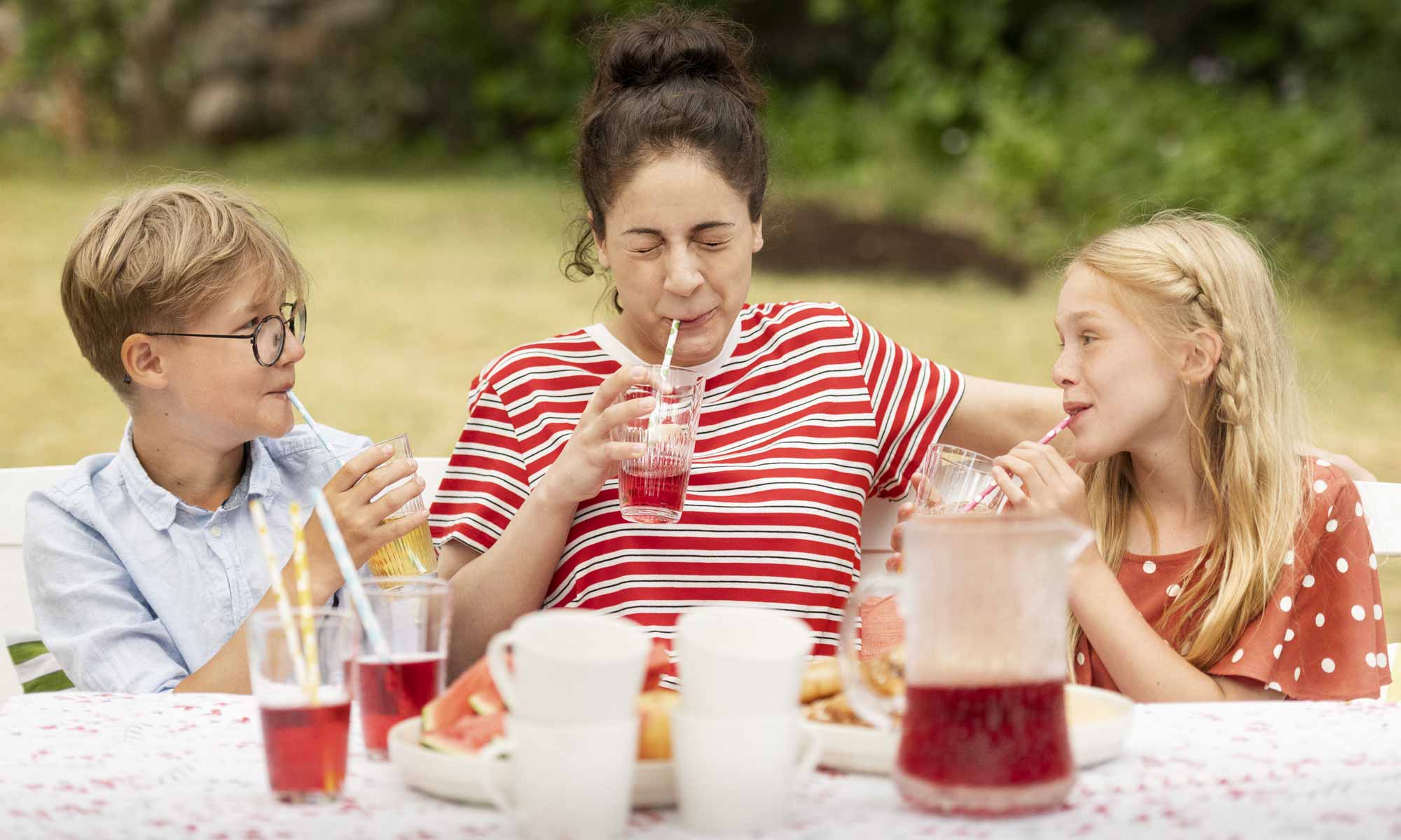 Mamma och två barn sitter ute i trädgården framför ett dukat fikabord och dricker saft 
